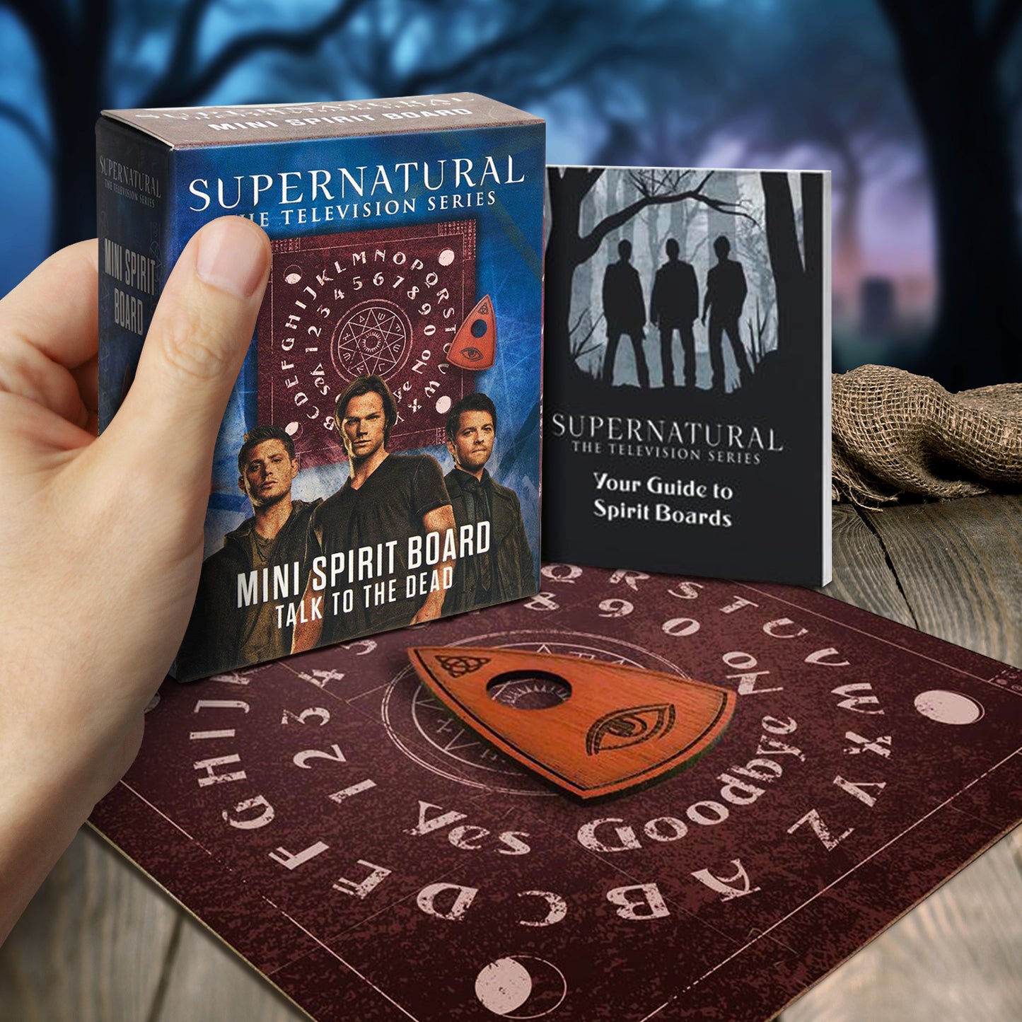 A brown ouija board on a wooden table, with a dark forest in the background. In the center is a wooden planchette. Behind the board is a book with instructions, and a cardboard box for the set. The box depicts Sam, Dean, and Castiel. A model's hand is holding the box.