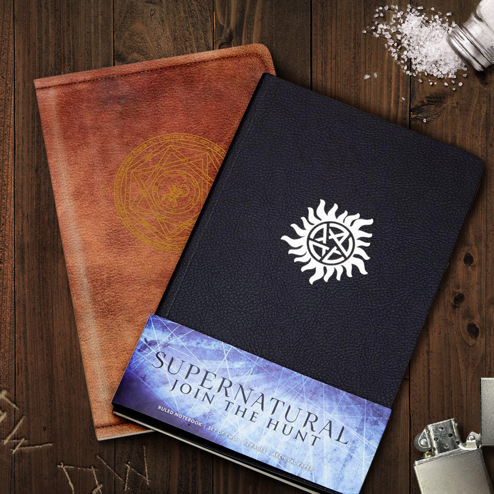 Two small journals on a wooden table. The front journal has a black cover, and features the Anti-Possession symbol in silver. The back journal is brown, with the Devil's Trap symbol in yellow. Across the bottom is a blue and white banner with black text that says "Supernatural: join the hunt." Next to the journals is a smal pile of salt, a metal lighter, and the initials SW and DW carved into the table.