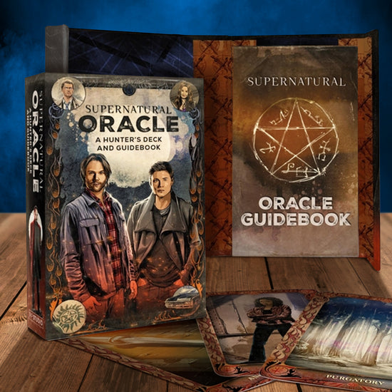 An oracle card set on a wooden table. The box has a painted depiction of Sam and Dean Winchester on the front. Next to the box are a few of the oracle cards from the set. In the background is the oracle guidebook, which has a Devil's Trap drawn on the front.