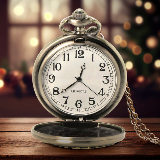 A silver pocket watch on a wooden table, open to show the white clock inside. Behind the watch is a faded and blurry christmas tree and window with christmas lights. A silver chain is attached to the top of the watch.