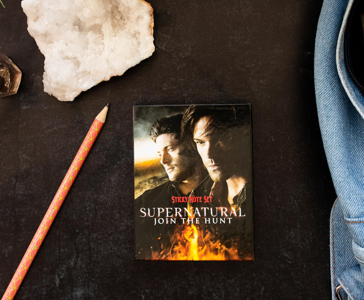 A sticky note box set featuring Sam and Dean Winchester on the front. Red and white text says Supernatural, Join The Hunt, sticky note set. The set is on a table next to a pencilm salt and a denim jacket.