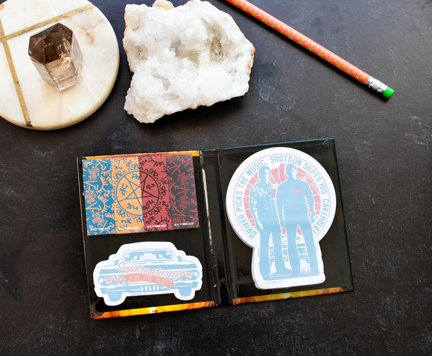 A collection of Supernatural-themed sticky notes, featuring Sam and Dean, Baby, and the Devils Trap symbol. Next to the notes are a pencil, a salt rock, and a crystal on a marble coaster.