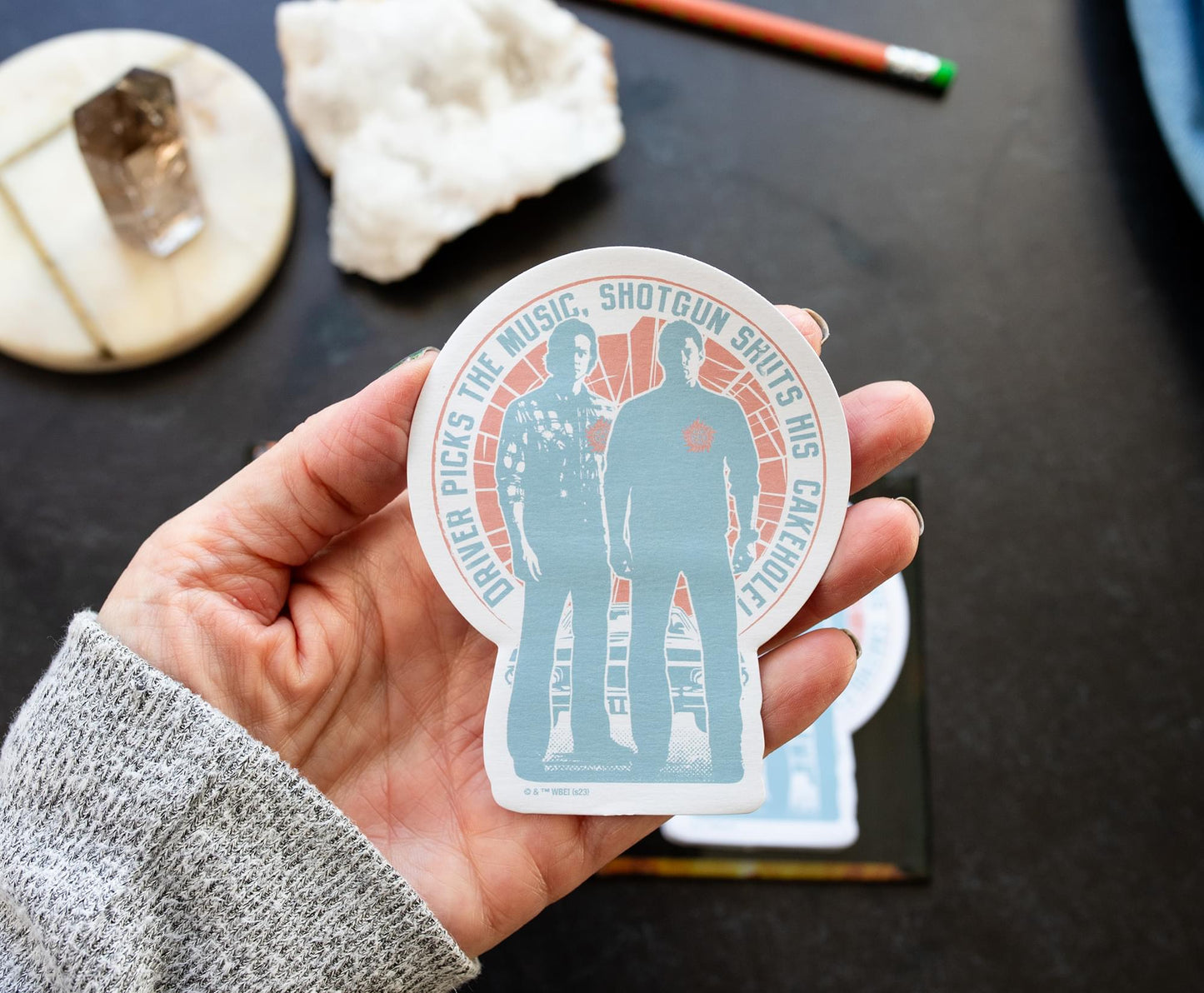 A model's hand holding a Supernatural-themed sticky note, featuring Sam and Dean.