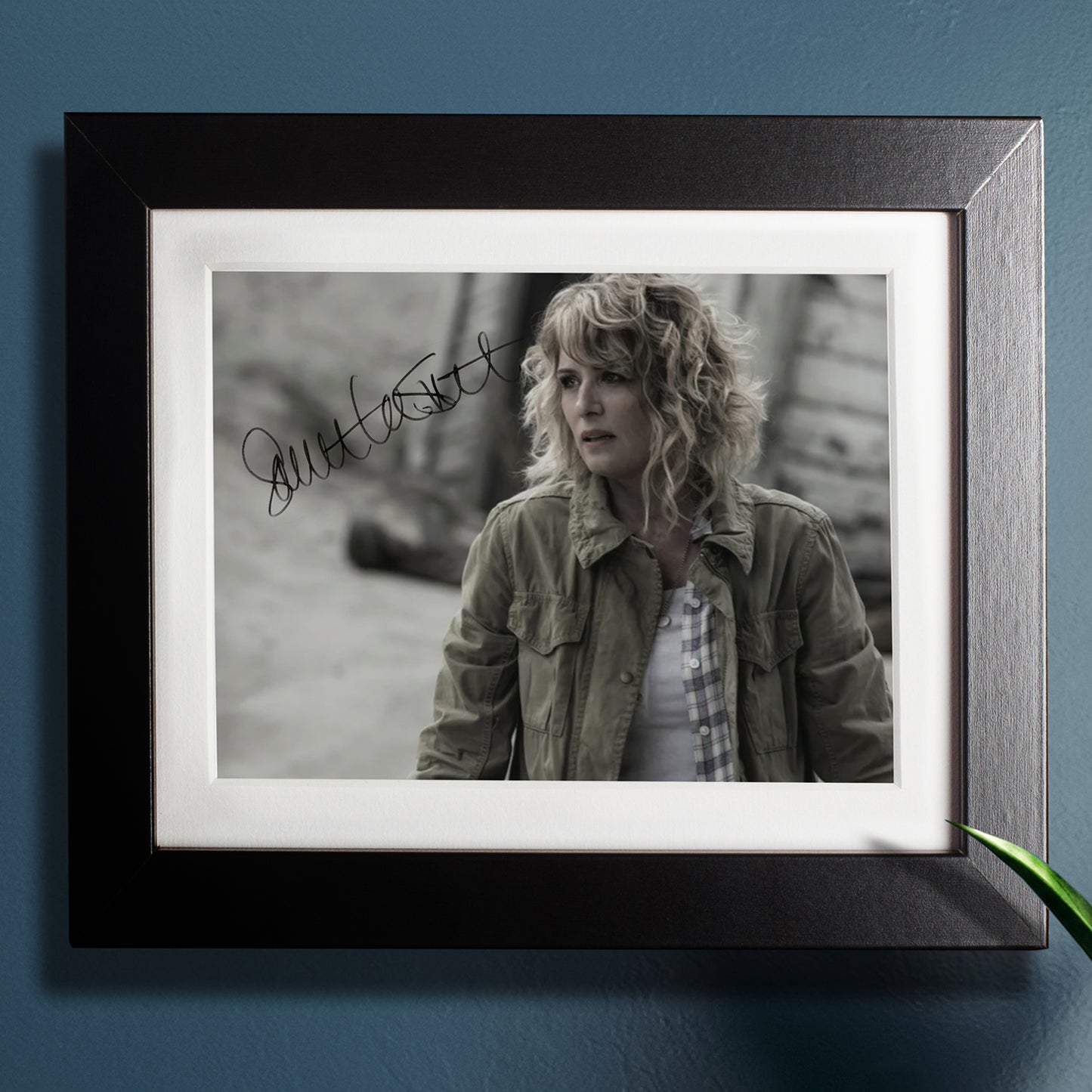 A black framed photograph against a dark blue wall. The photograph depicts actor Samantha Smith, dressed as the character Mary Winchester from the series "Supernatural." Samantha is wearing a plaid shirt under a green jacket. Behind her is a small cabin. In the top left corner is Samantha's autograph in black ink.