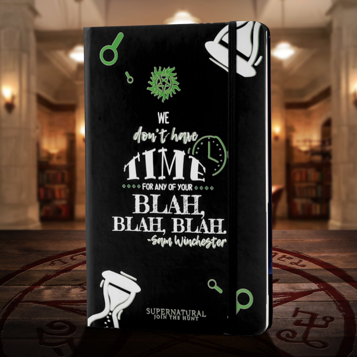 A black journal standing on a wooden table with a red devil's trap symbol on it. The cover has drawings of white hourglasses in the corners, and a green anti-possession symbol in the center. White text says "we don't have time for any of your blah, blah, blah. Sam winchester." Behind the book is the bunker from the TV series Supernatural