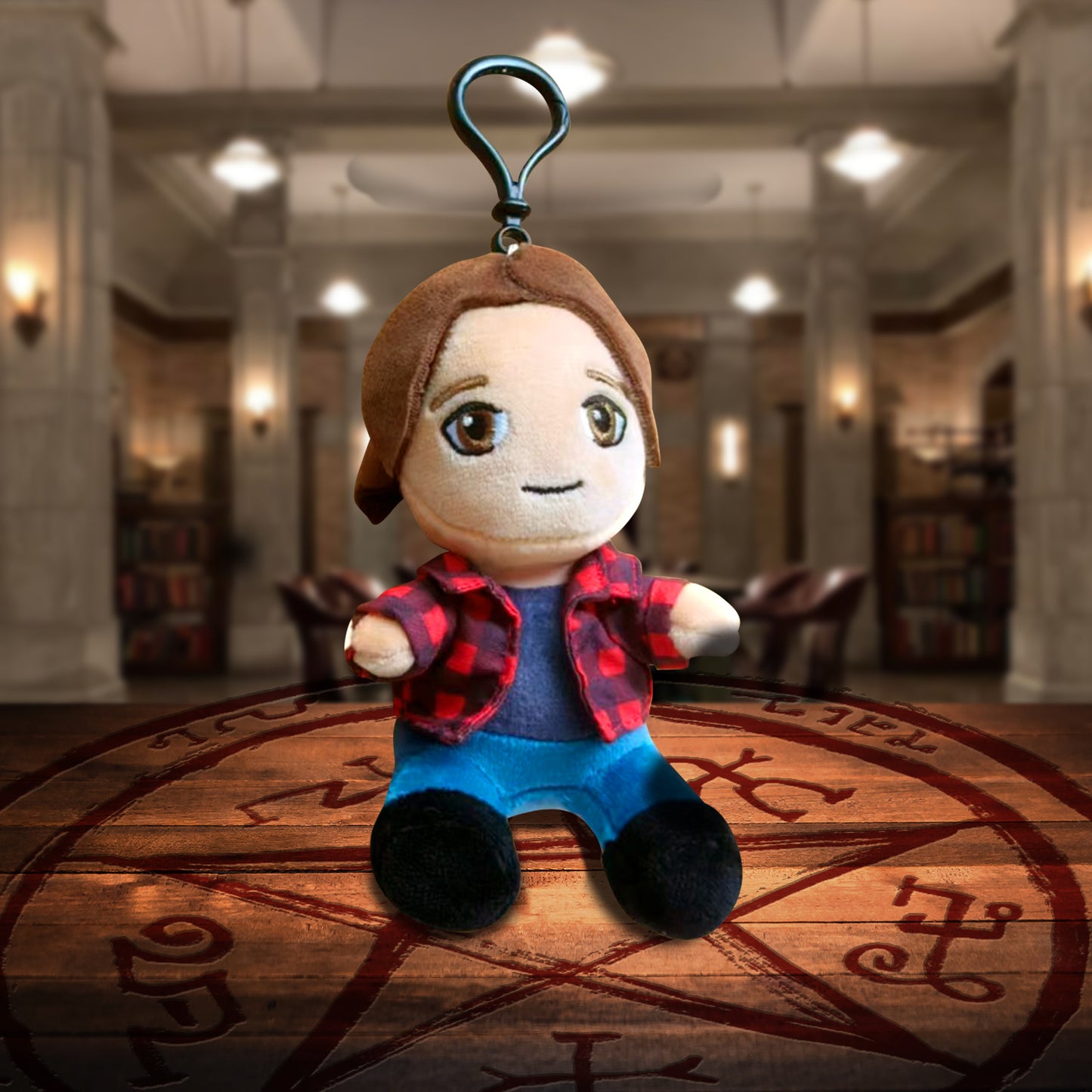 A plush keychain depicting Sam Winchester from Supernatural. Beneath the keychain is a Devil's Trap on a wooden table. In the background is The Bunker.