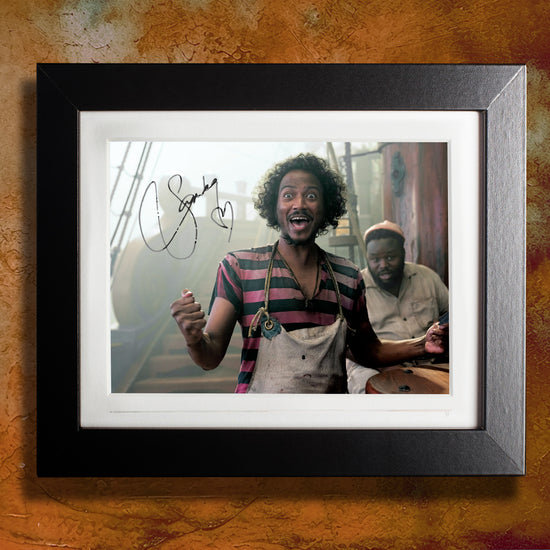 A black framed photograph against a brown wall. The photograph depicts actor Samba Schutte, dressed as the character Roach from the series "Our Flag Means Death." Samba is in a pink and black striped shirt, with a white apron on. Behind him is a pirate ship. On the top left side is Samba's autograph in black ink.