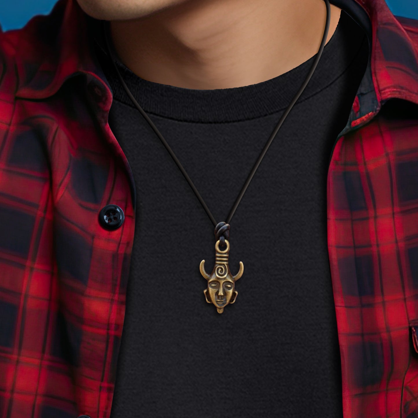 An image of a model wearing a necklace with a brass pendant, sitting against a black tee shirt and a red plaid flannel shirt. The pendant is shaped like the Samulet amulet.