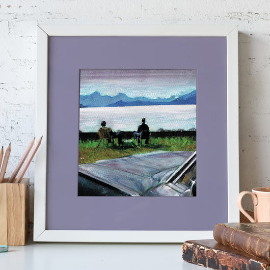 A white-framed lithograph against a white stone wall. The lithograph depicts Sam and Dean Winchester sitting in front of a lake, with a black Impala in the foreground. In front of the lithopgrah is a pencil holder and a pair of leather-bound books.