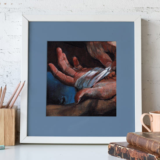 A framed lithograph against a white wall. The lithograph depicts a pair of hands wrapped in white bandages. In front of the lithograph is a wooden pencil holder, a dna pair of brown leather-bound books