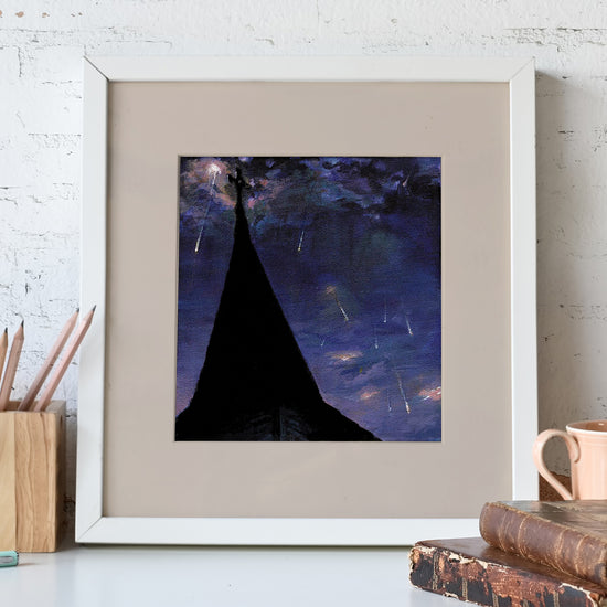 A white-framed lithograph against a white stone wall. The lithograph depicts a church steeple in silhouette, against a purple nighttime sky. In front of the lithopgrah is a pencil holder and a pair of leather-bound books.