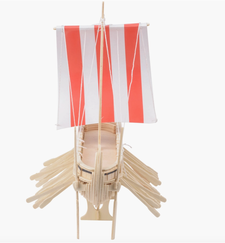Front view of a miniature wooden Viking ship with a read and white sail, set against a white background. The ship has many oars sticking out the sides, and miniature wooden shields line the edge, with drawings of axes and dragon heads on each.