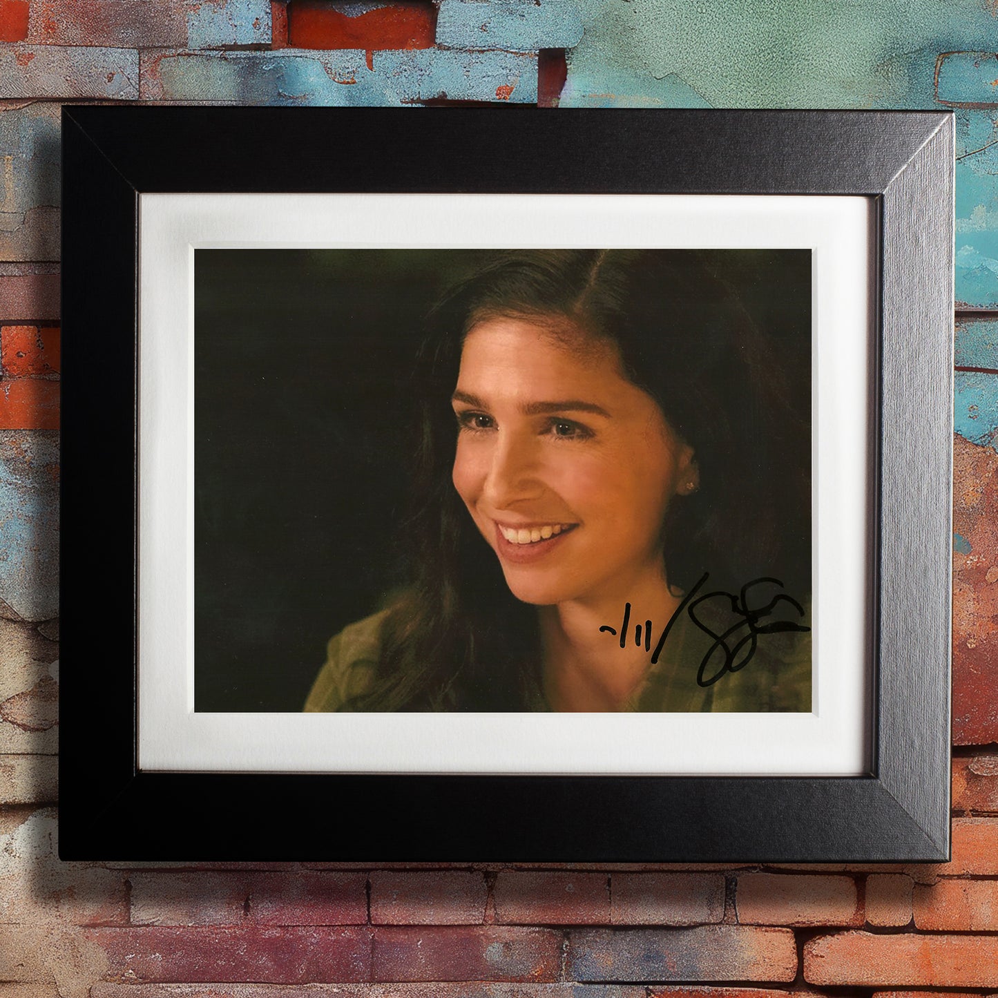 A black framed photograph against a colorful brick wall. The photograph depicts actor Shoshannah Stern, dressed as the character Eileen Leahy from the series "Supernatural." Shoshannah is wearing a green jacket, with a bright smile on her face. In the bottom right corner is Shoshannah's autograph in black ink.