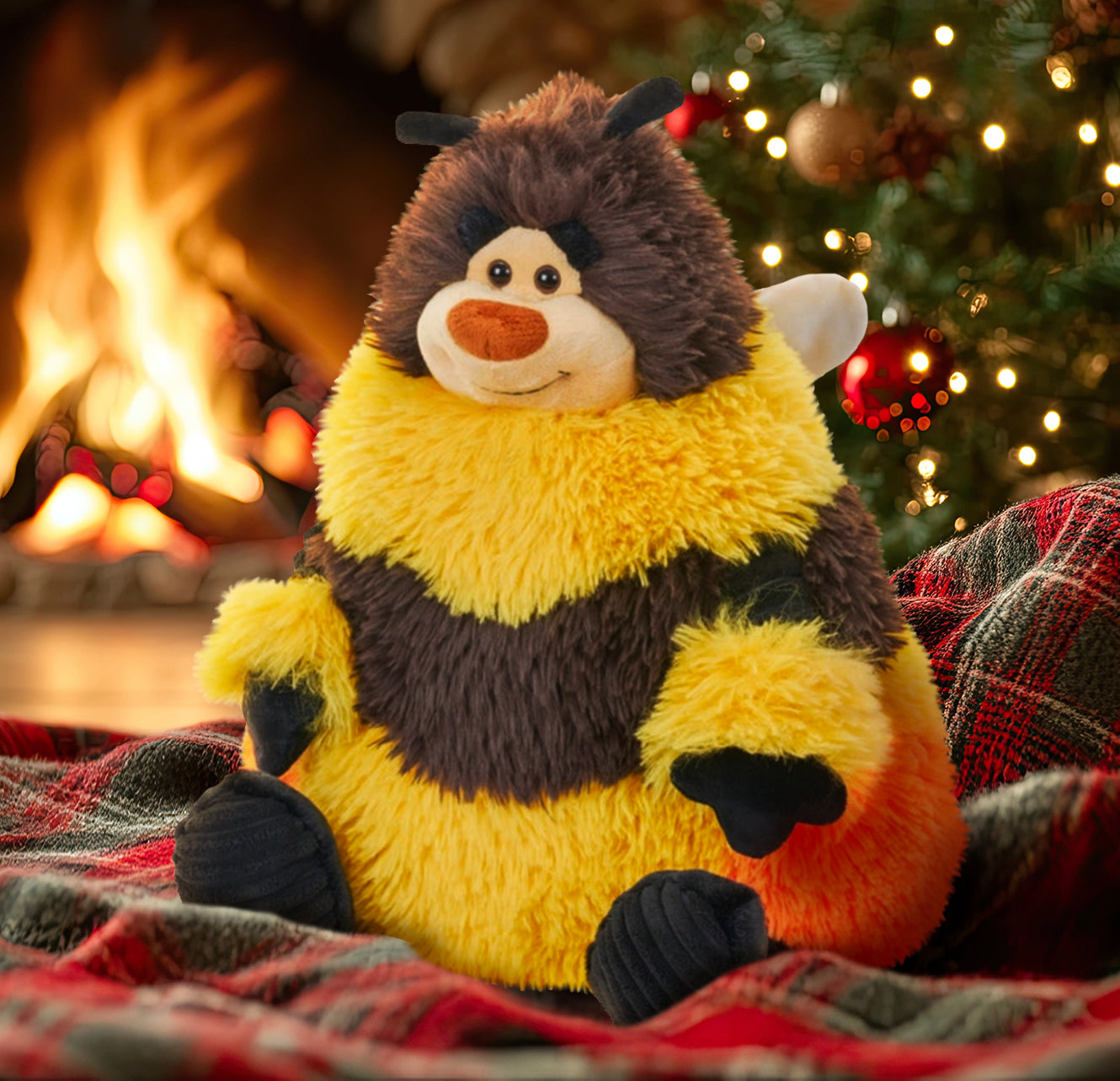 A bee plushie sitting on a red and green plaid blanket. The bee has yellow and brown striped, with white wings on his back. At his bottom is a patch of orange, to represent pollen. The bee has black hands and feet, and a smile on his face. Behind the bee is a Christmas tree with white lights, next to a roaring fireplace.