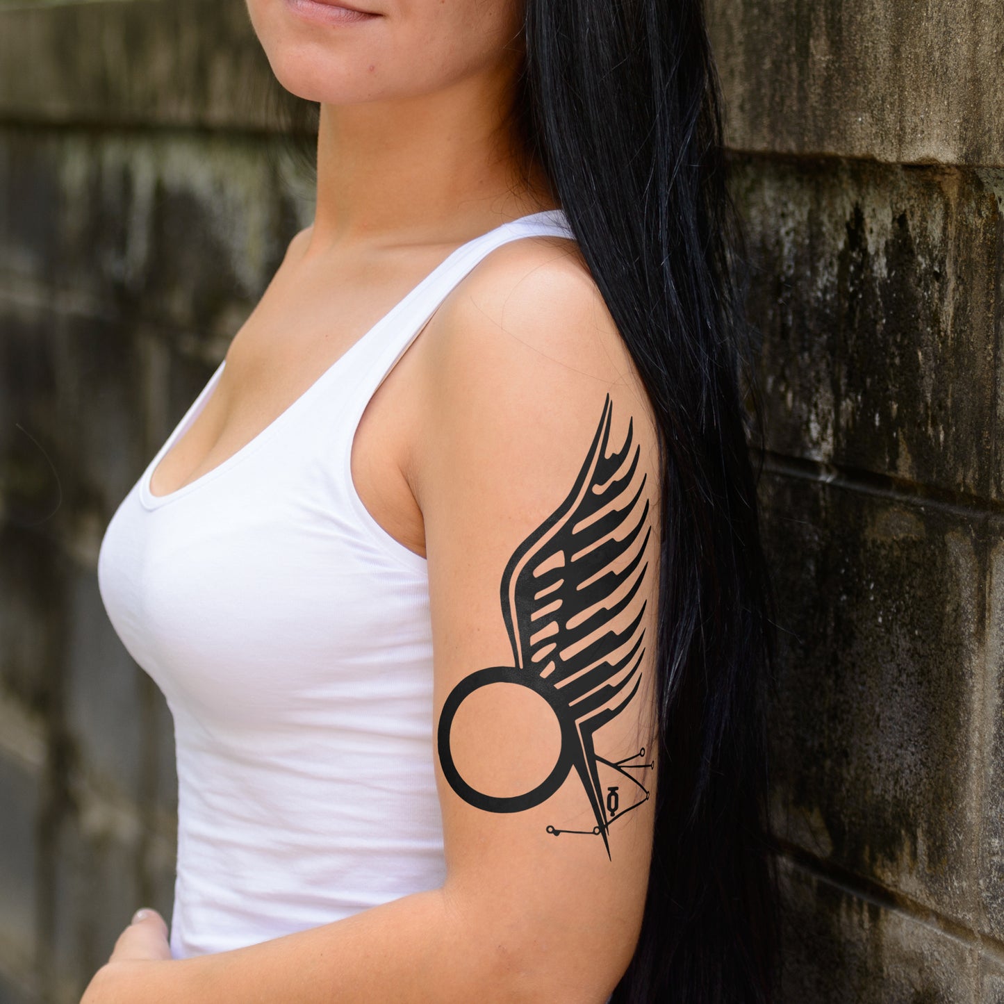 A female model in a white tank top, standing against a concrete wall. On her arm is a black tattoo depicting a circle with an angel wing on one side.