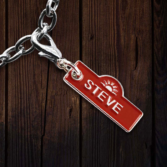 A red name tag with the name STEVE printed in the center in white text, and a white rising sun motif above the name. Behind the charm is a dark wooden table.