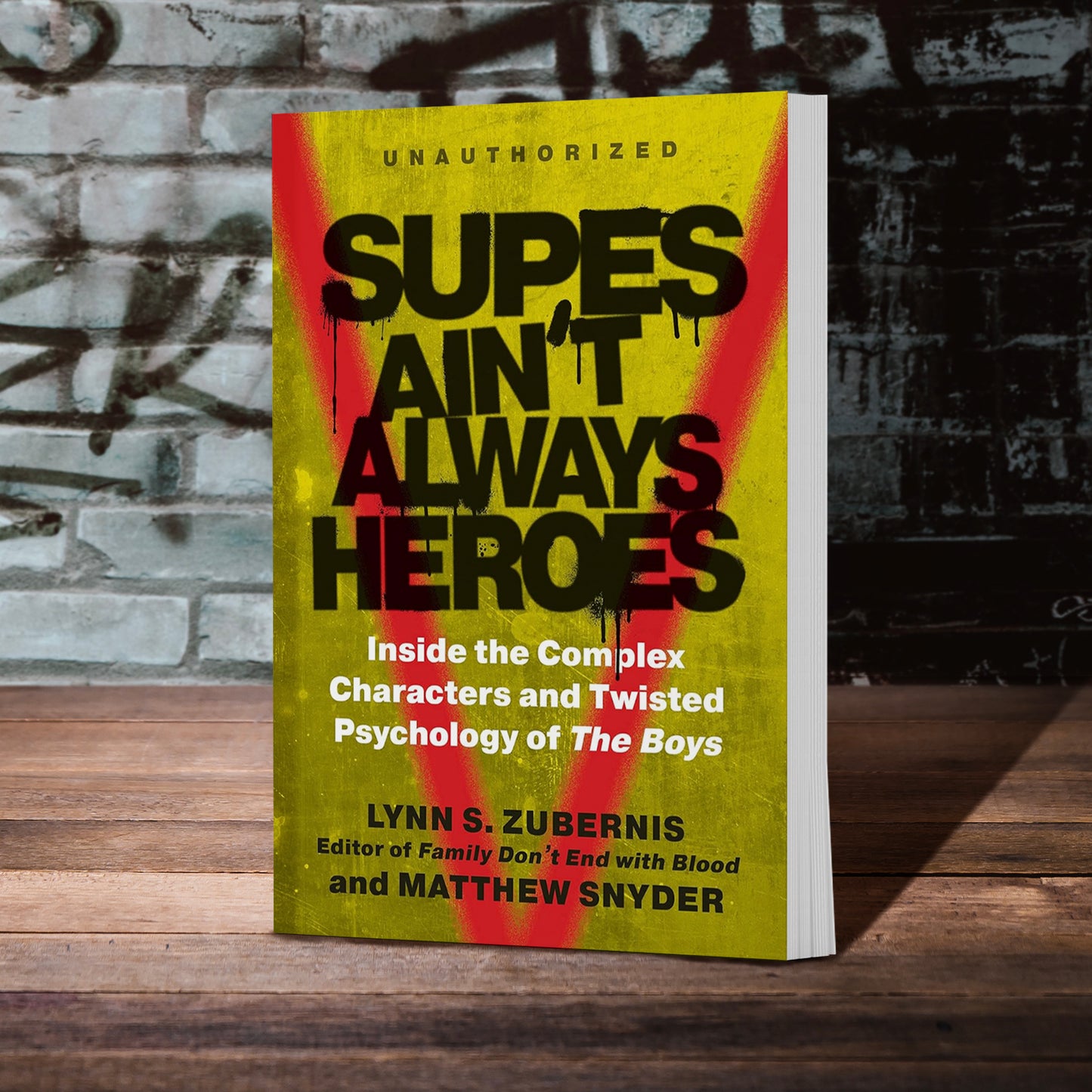 An image of a grainy yellow book on a wooden table. At the top in black text is "Unauthorized: Supes Ain't Always Heroes." Under the title in white text is "Inside the complex characters and twisted psychology of The Boys." A red spray-painted V is under the text in the background. Behind the book is a grey brick wall covered in graffiti.