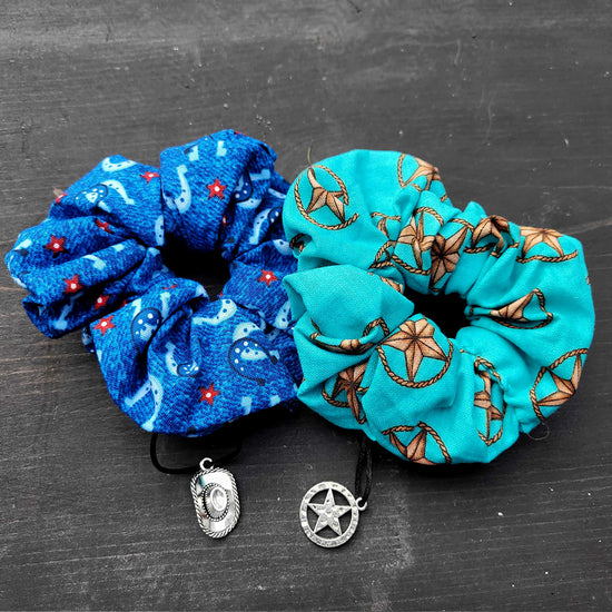 Two blue scrunchies side by side on a black table. The left is medium blue with a pattern of horseshoes and red starts. The right is light blue with a pattern of gold star badges.