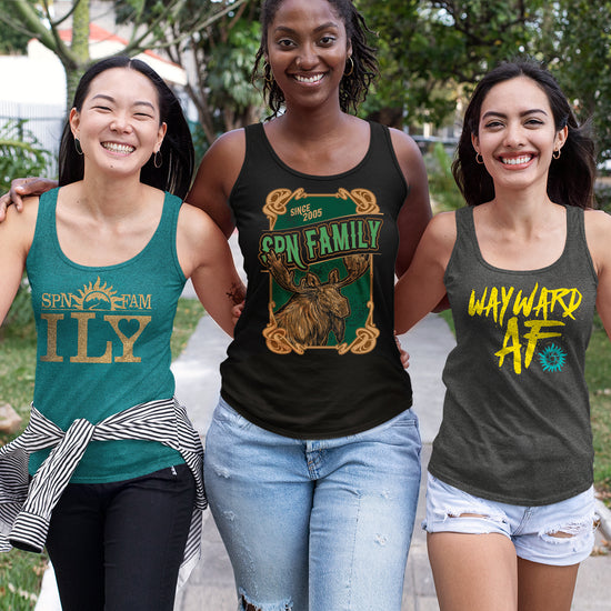 Three female models wearing tank tops, with a park in the background. The left model is in a teal tank, with "SPN FAM ILY" in gold text. The moddle model is in a black tank, with a cartoon drawiing of a moose in a green frame, and "SPN Family" in green text. The right model is in a grey tank, with "Wayward AF" in yellow text.