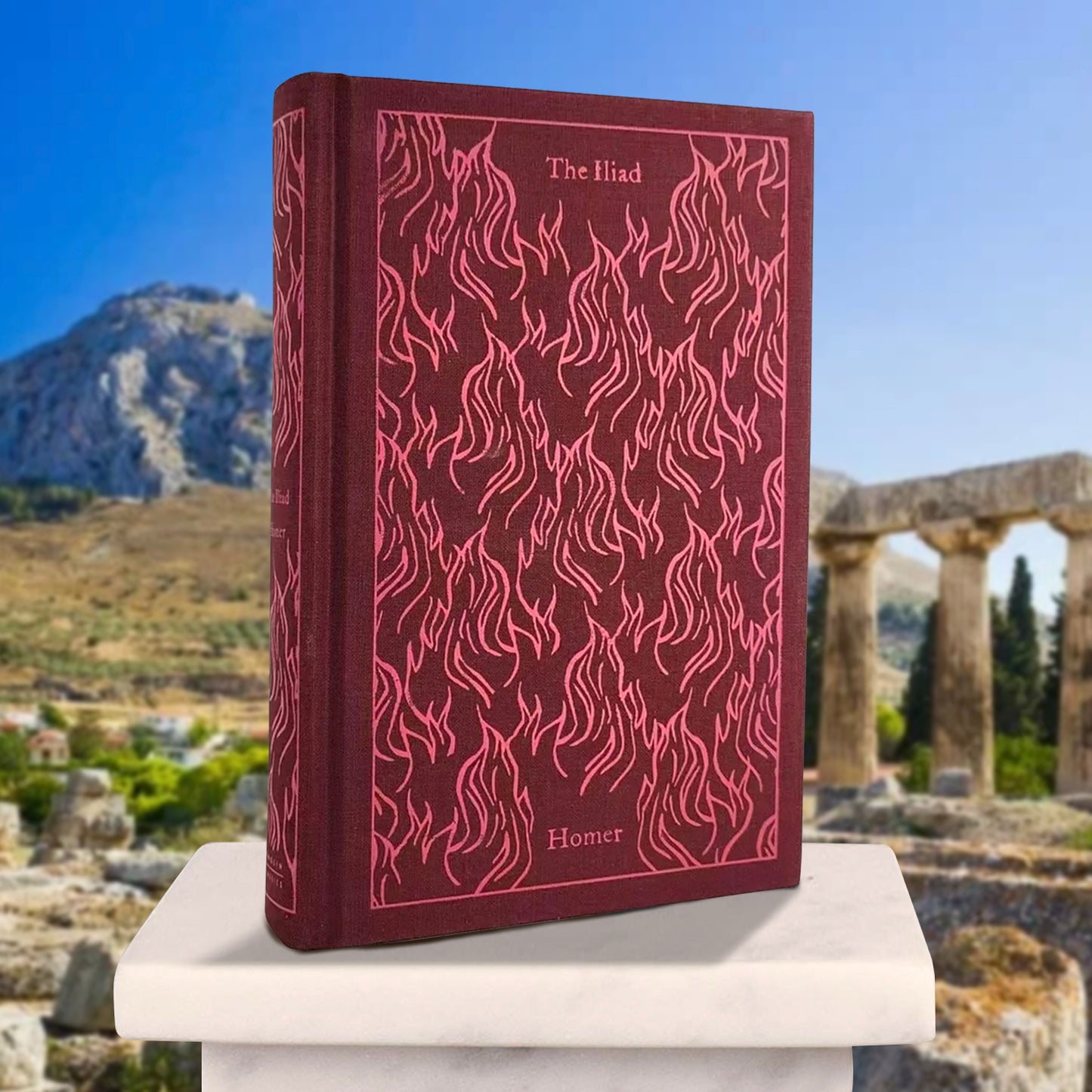 A dark maroon book on a white marble pillar, with mountains and ruins in the background. The front cover has light red drawings of flames across it. Light red text at the top and bottom says "The Iliad, Homer"