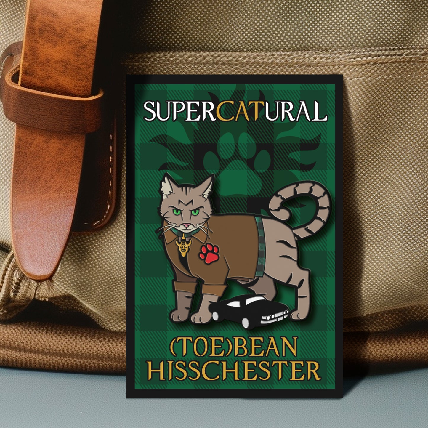 An enamel pin of a grey-brown tabby cat with bright green eyes. The cat has a mischievous expression and is wearing a brown jacket with the edge of a green flannel showing underneath, as well as a 'Samulet' collar charm. It has a red paw print on one shoulder and a 'plush toy' Impala between its feets. The pin backing reads: "SuperCATural - (Toe)Bean Hisschester)"
