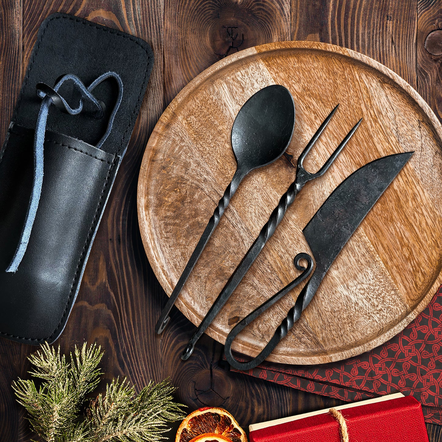 A spoon, form, and knife on a wooden plate, all made of black iron, on a wooden plate. Each has a decorative twist along the length of its shaft. The fork has two prongs, and the knife's handle folds back on itself. Next to the plate is a black leather holder for the cutlery. Below the plate is a sprig from a green tree, a black and red napkin, dried fruit, and a red box.