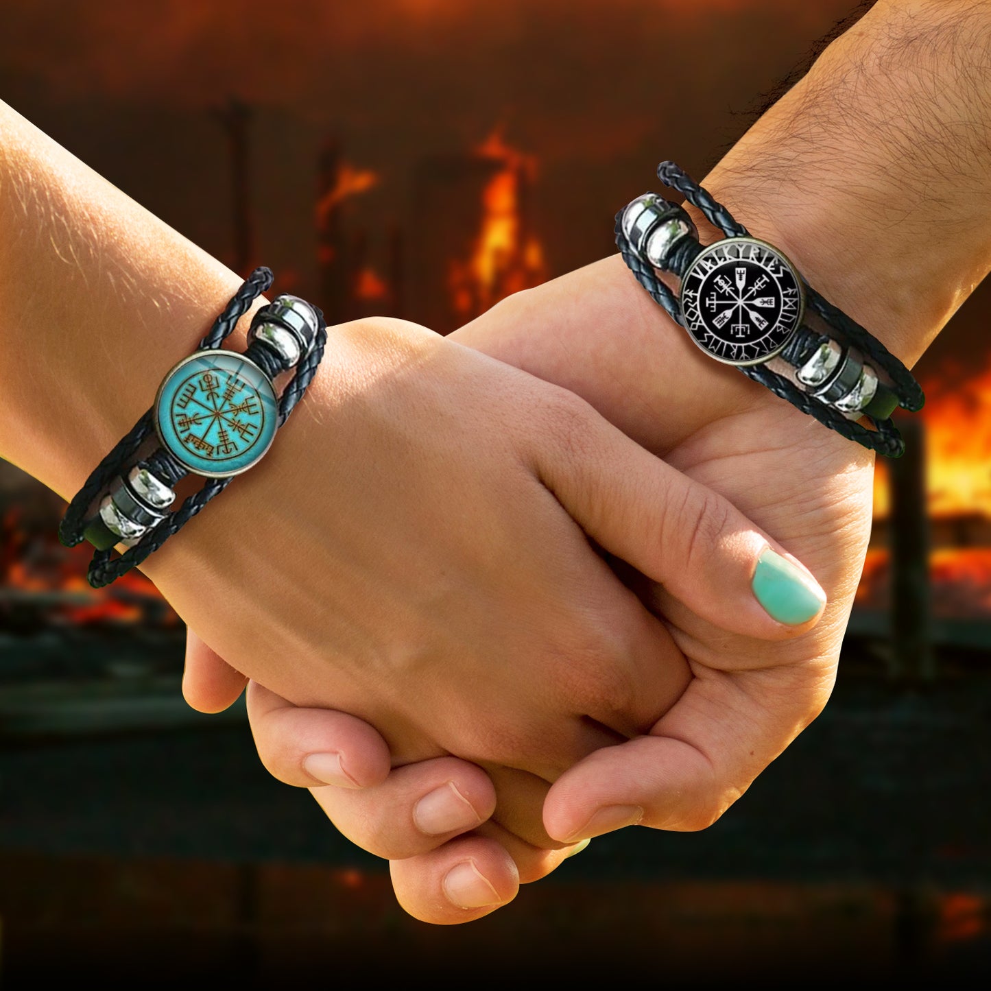 Close up of two models's hands, clasped together. Each model is wearing a black braided bracelet. The left bracelet has a blue and gold round charm, the right bracelet has a black and silver charm. Each charm depicts Nordic runes in the shape of a compass. Behind the hands is a blurred fiery village.