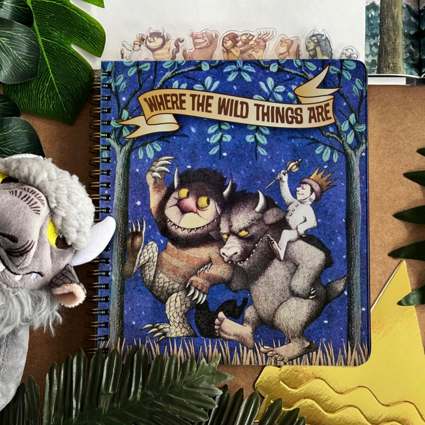 A blue journal depicting Max and two monsters from Where the Wild Things Are. Two monsters are carrying Max on their shoulders. At the top is a banner across two trees with text saying where the wild things are. At the top of the journal are tabs depicting characters from the book. Next to the journal is a goat puppet and plants
