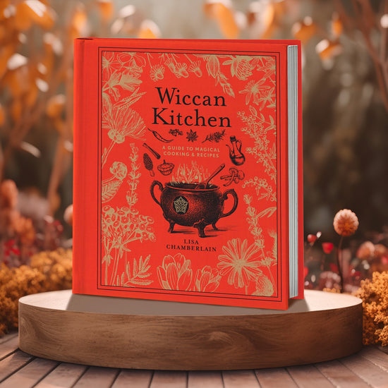 A red and black book on a wooden cutting board. Scattered across the book cover are drawings of various herbs, in gold ink. At the center is a black drawing of a witch's cauldron and various witchy ingredients. At the top is black and gold text saying "Wiccan Kitchen: a guide to magical cooking & recipes." Fall-colored leaves are behind the book.