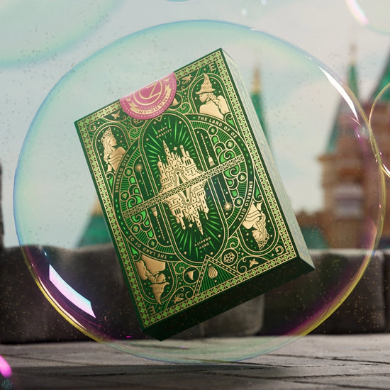 A green and gold box of playing cards suspended in a soap bubble. The box depicts images from the movie Wicked in green and gold. In the background is a colorful castle.