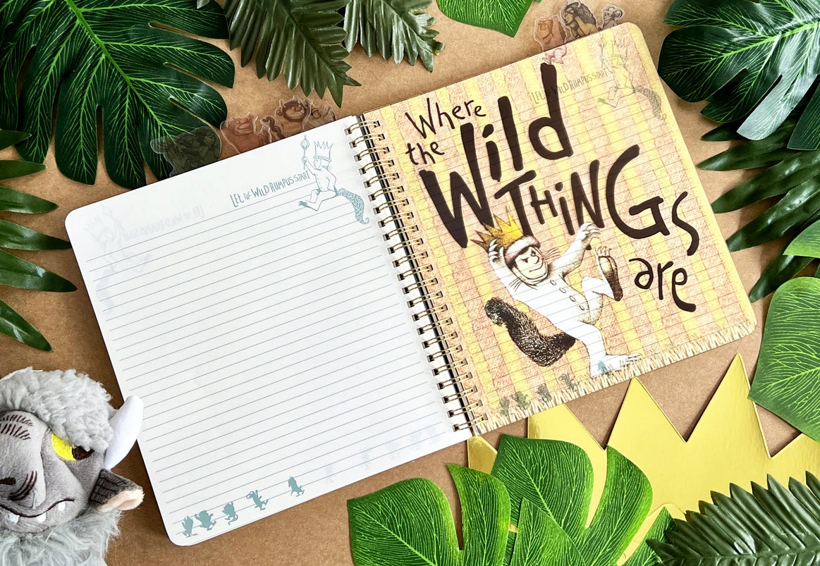 A page from the book showing Max on a yellow striped background, carrying a scepter. Black text says "where the wild things are." The journal is on a table surrounded by leaves and a goat puppet.