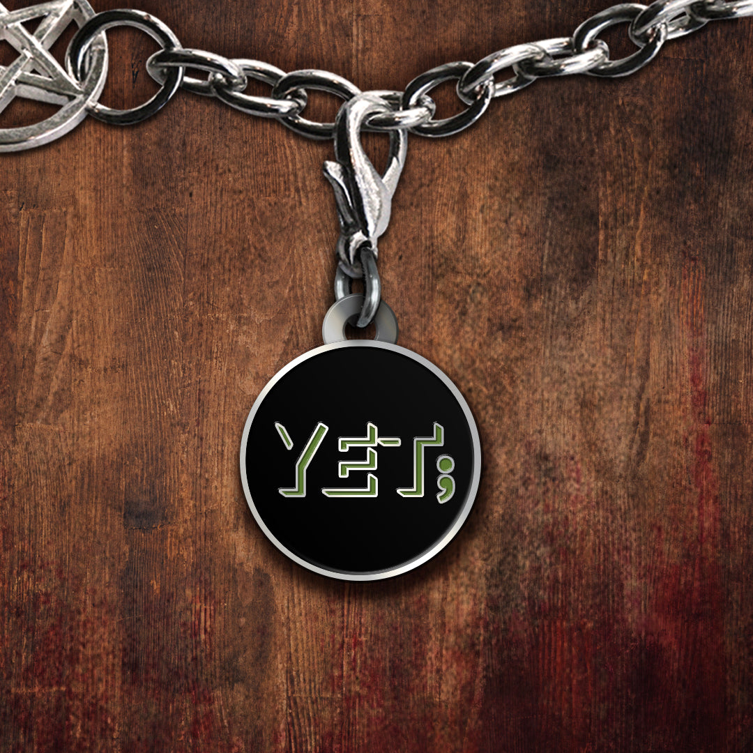 A silver-toned black charm featuring the word 'YET;' in stylized green letters, displayed on a dark wood background.