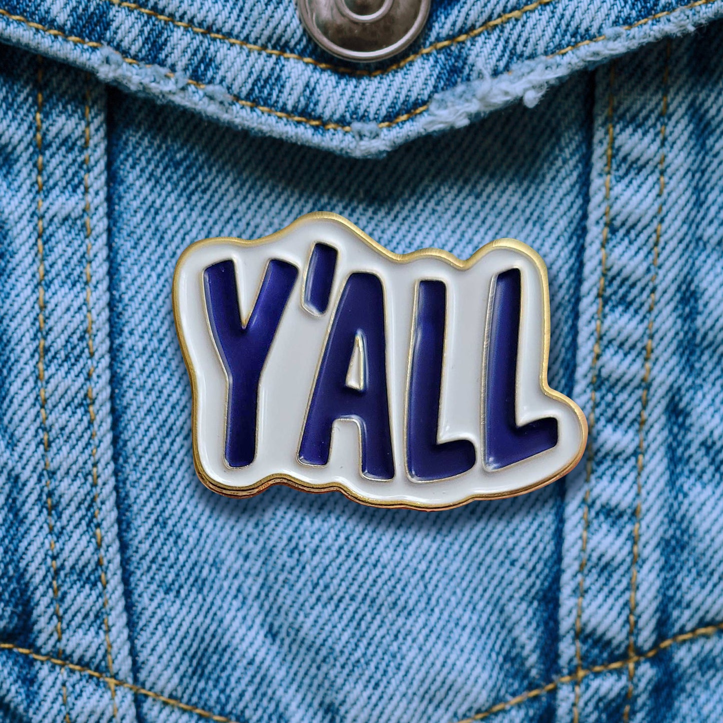 An enamel pin attached to a blue denim pocket. The pin is white, with blue text saying "y'all."