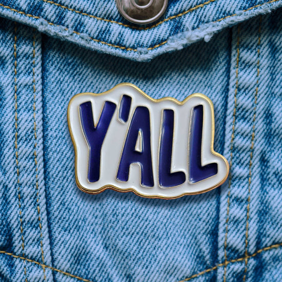 An enamel pin attached to a blue denim pocket. The pin is white, with blue text saying "y'all."