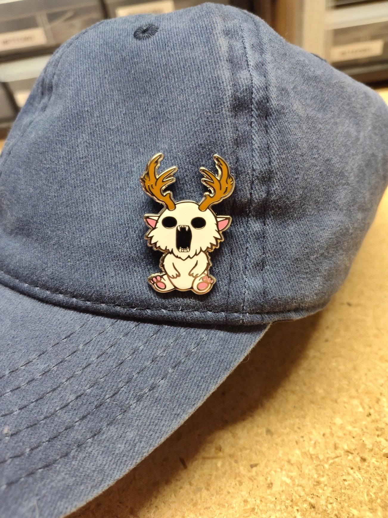 A metal pin depicting a white creature with antlers. The pin is on a white backing card, with black text saying "Mountain's misfits. We're all weird sometimes." The pin is attached to a black baseball hat.
