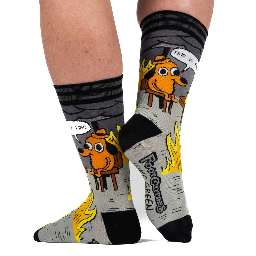 A model's feet wearing a pair of grey and back socks. One sock has a cartoon of a smiling brown dog wearing a hat, sitting in a chair in front of burning flames. Above him is a caption saying "This is fine." The other sock has a brown wooden table with a coffee cup, and a roaring flame underneath it. The cuff and toes of the socks are black.