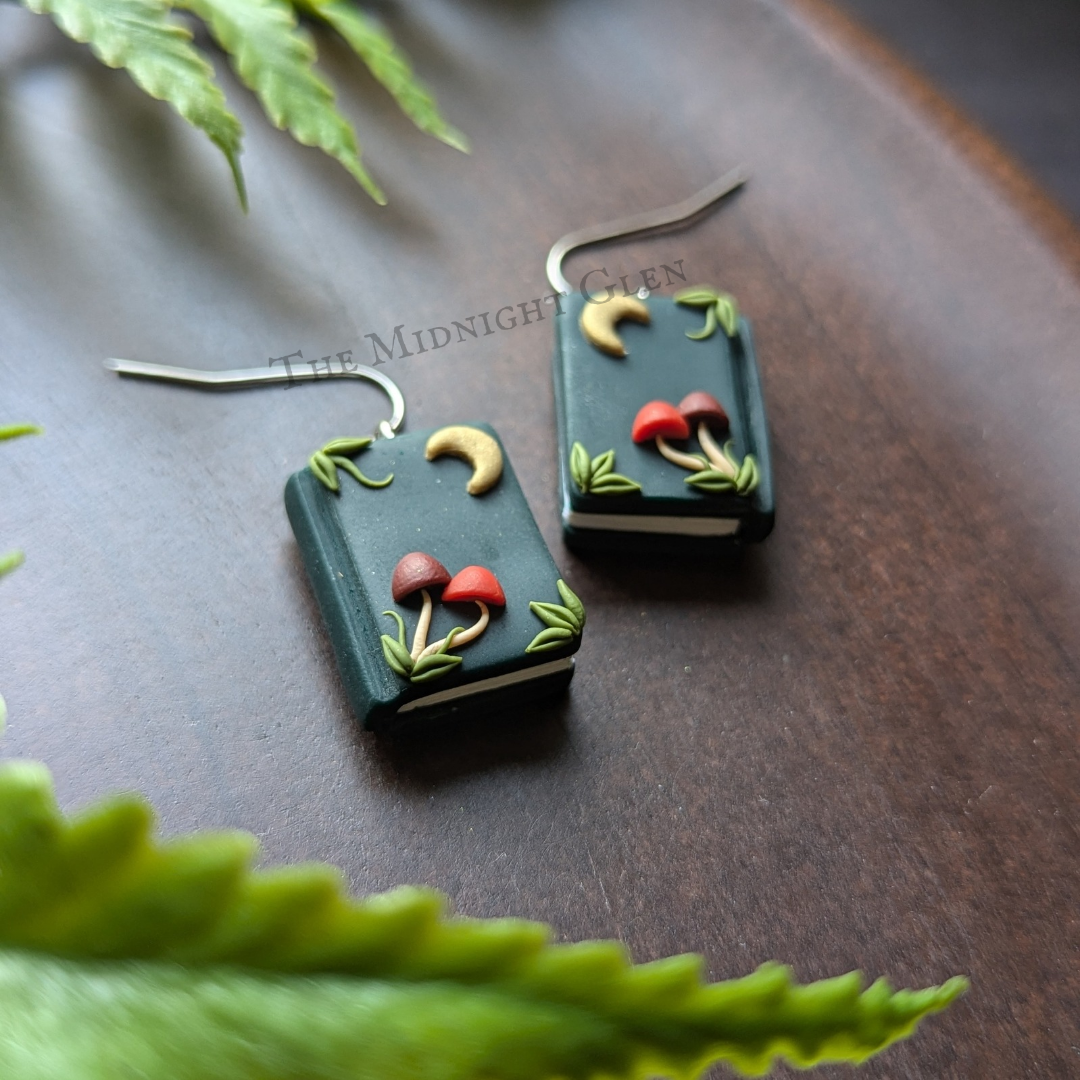 A pair of dangle earrings shaped like books, with tiny moons and mushrooms on the front.