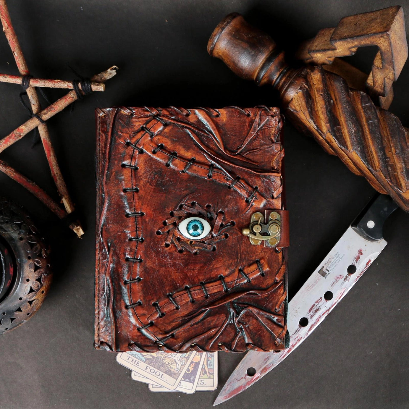 A leather journal against a grey table. The cover is styled to appear stitched together with a blue eyeball in the center. Around the journal are tarot cards, a red-stained knife and wooden trinkets.