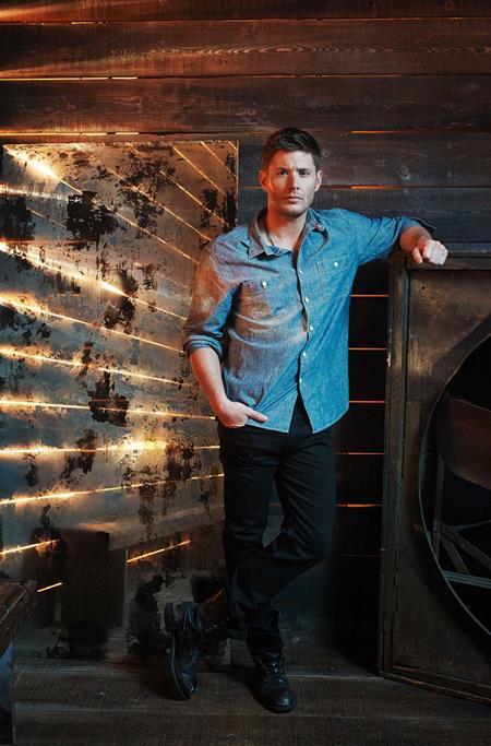 A man wearing a blue denim button-up shirt and black pants stands in a rustic industrial setting with a dark wood wall and warm light strips.
