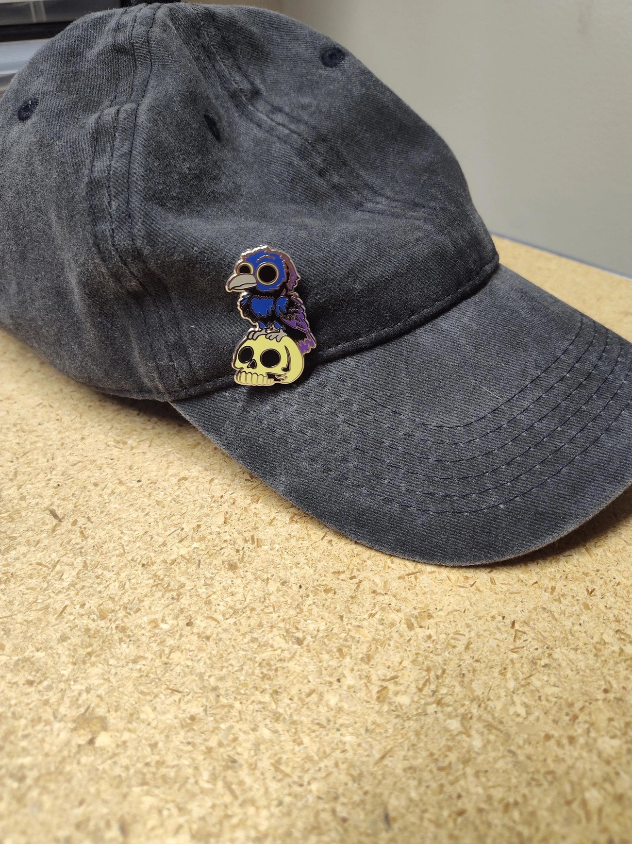 A metal pin depicting blue and grey crow, perched on a yellow skull. The pin is on a white backing card, with black text saying "Mountain's misfits. We're all weird sometimes." The pin is attached to a black baseball hat.