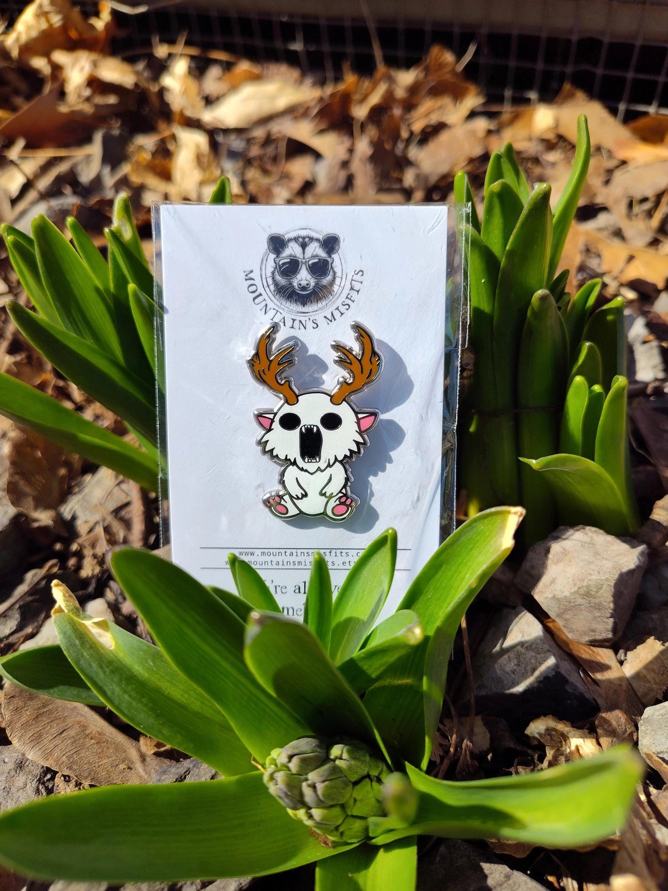 A metal pin depicting a white creature with antlers. The pin is on a white backing card, with black text saying "Mountain's misfits. We're all weird sometimes." The pin is leaning against green leaves and rocks.