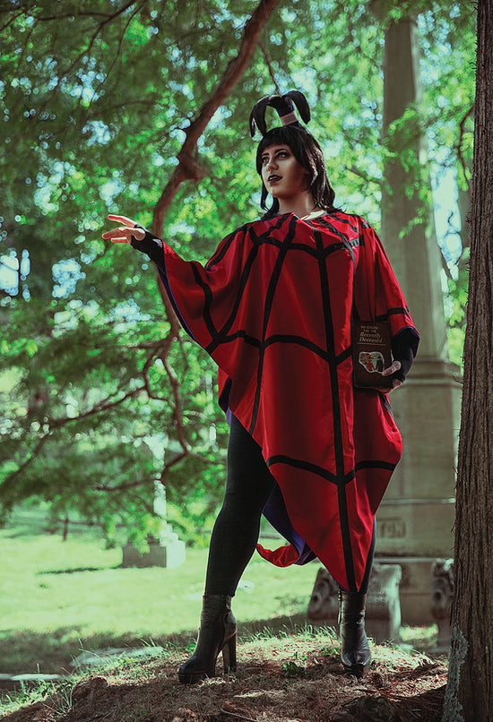 A cosplayer dressed as Lydia Deetz in a red spider-web cloak and holding a "Handbook for the Recently Deceased" 