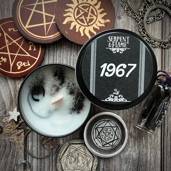 An opened candle tin on a grey wood table. The lid is black with white text saying "serpent & flame: 1967." Scaattered around the candle are various charms and trinkets with symbols from the SPN TV series, including the anti-possession symbol and the Devil's Trap.