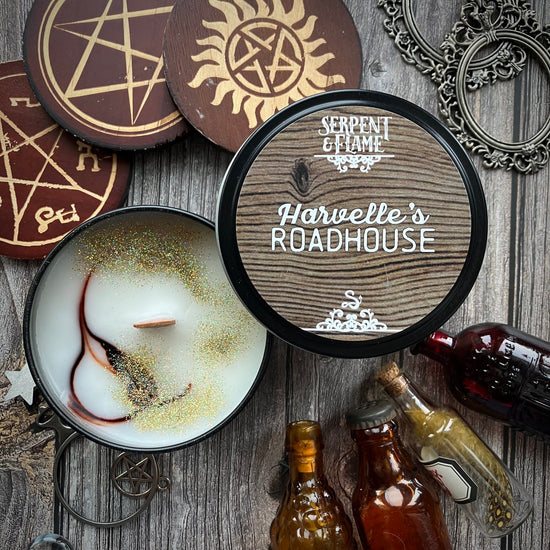 An opened candle tin on a grey wood table. The lid is brown with a wooden grain pattern. White text on the lid says "serpent & flame: Harville's Roadhouse." Scaattered around the candle are various charms and trinkets with symbols from the SPN TV series, including the anti-possession symbol and the Devil's Trap. Miniature beer and whiskey bottles sit on the table nex to the tin.