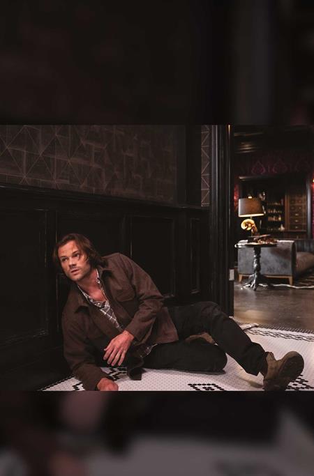 A man in a brown jacket and dark pants reclines on a patterned rug in a dimly lit interior room with dark wood paneling.