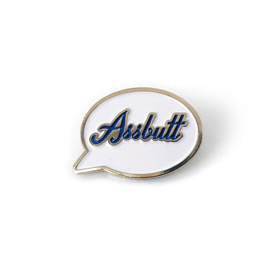 A silver pin with a white front in the shape of a speech bubble. The word "Assbutt" is written in blue script font.